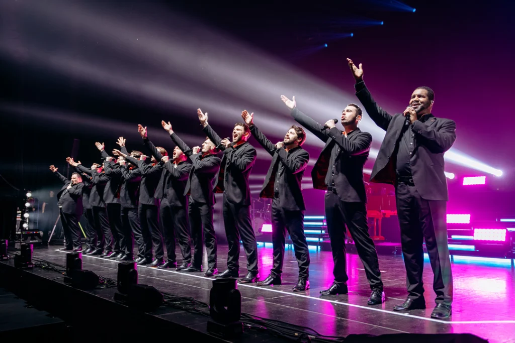 Die 12 Tenors auf der Bühne, in schwarzer Kleidung, mit erhobenen Armen während eines Konzerts.