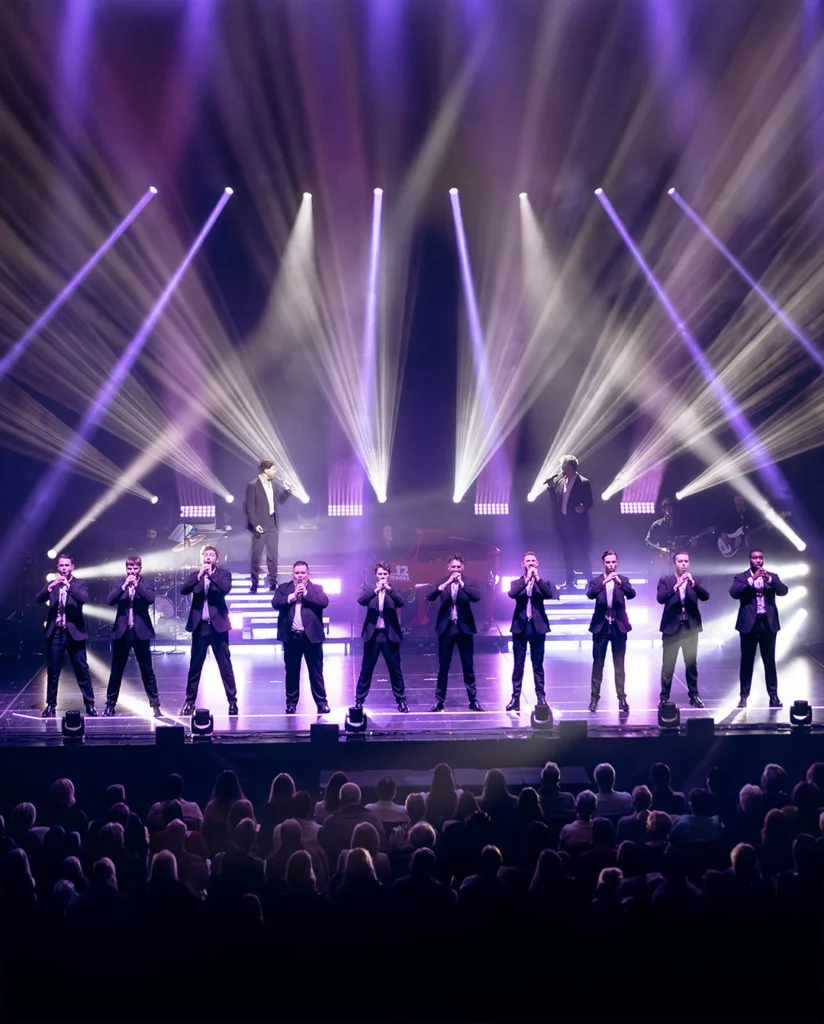 Teaserbild zu The 12 Tenors mit zehn Sängern auf der Bühne in Anzügen vor einem beleuchteten Hintergrund.