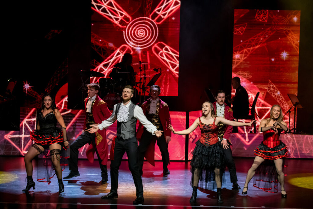 Teaserbild zu Moulin Rouge mit Tänzern in auffälligen Kostümen auf der Bühne.