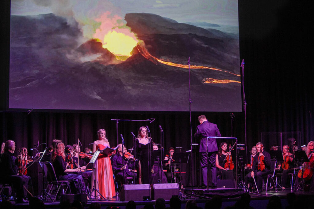 Teaserbild zu Der Herr der Ringe mit einem Orchester und einer Vulkanprojektion im Hintergrund.