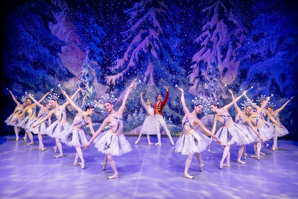 Tanzszene aus Der Nussknacker mit Balletttänzerinnen in weißen Kostümen vor einer winterlichen Kulisse.