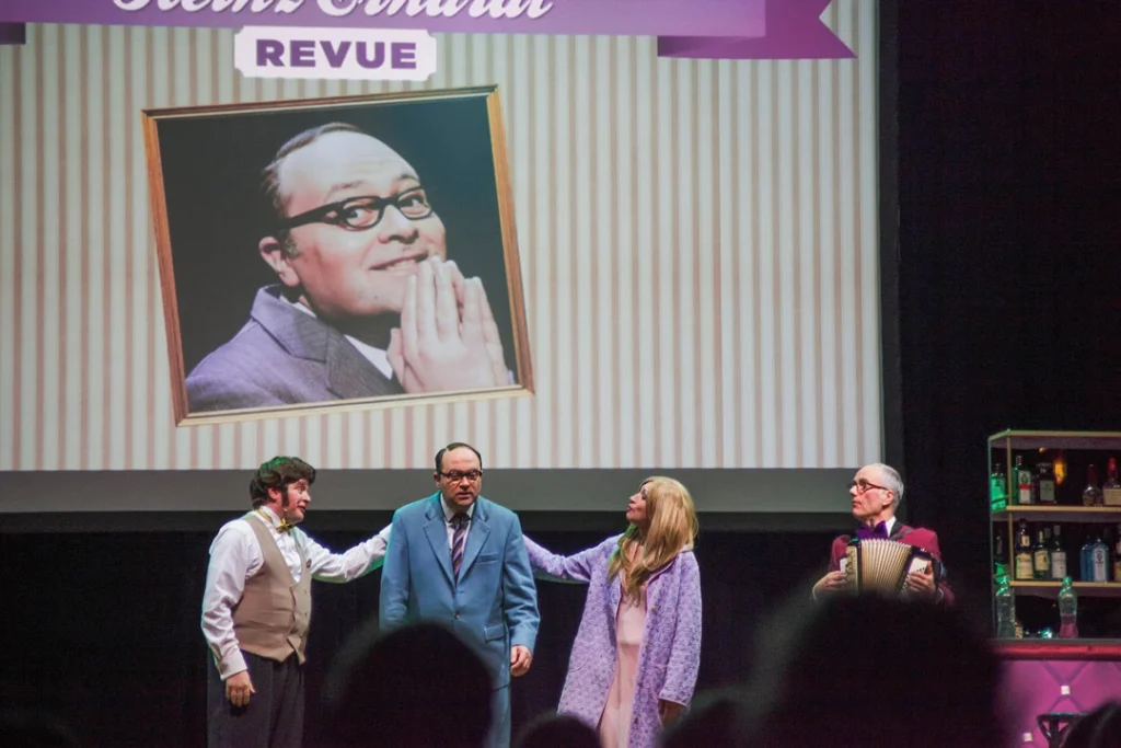 Teaserbild zu Die Neue Heinz Erhardt Revue mit vier Darstellern auf der Bühne und einem Bild von Heinz Erhardt im Hintergrund