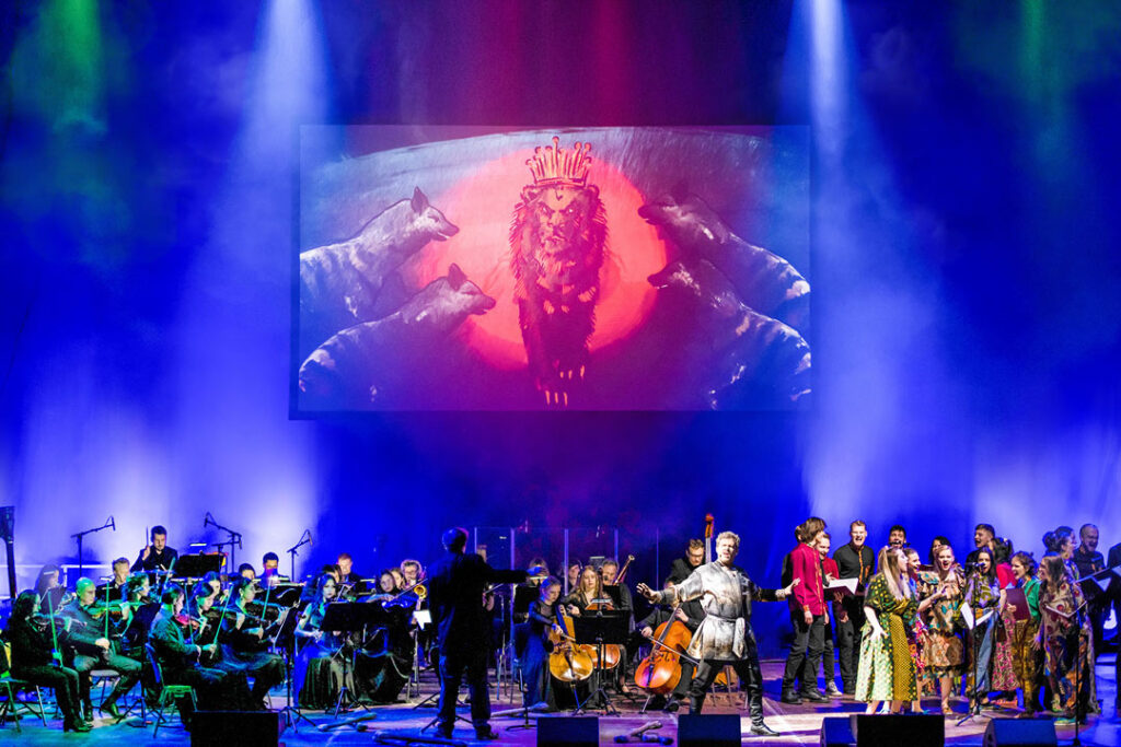 Teaserbild zu Der König der Löwen mit Orchester und Darstellern auf der Bühne vor einem großen Hintergrundbild.
