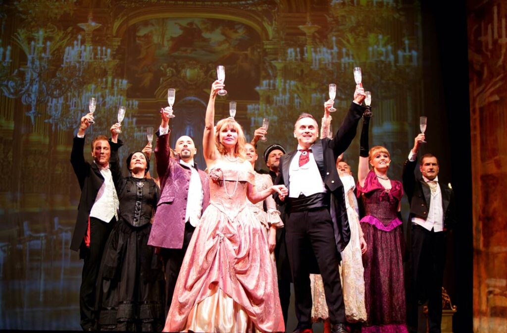 Teaserbild zu Das Phantom der Oper mit Darstellern in festlicher Kleidung, die Gläser heben.
