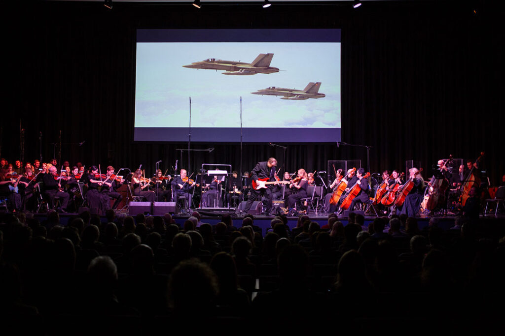 Konzertszene mit Orchester und Flugzeugen im Hintergrund während eines Hans Zimmer Events.