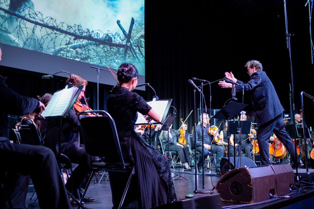 Orchesteraufführung mit Dirigent und Musikern auf der Bühne, im Hintergrund eine Projektion.