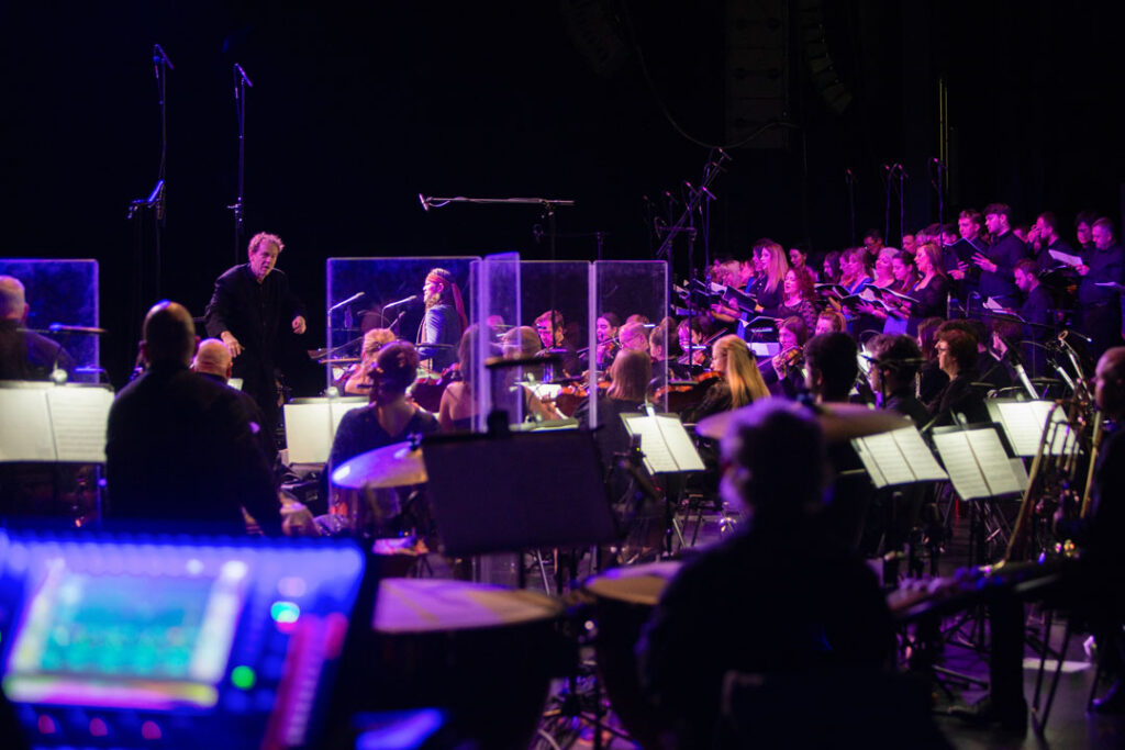 Orchesteraufführung mit Dirigent und Chor im Hintergrund.