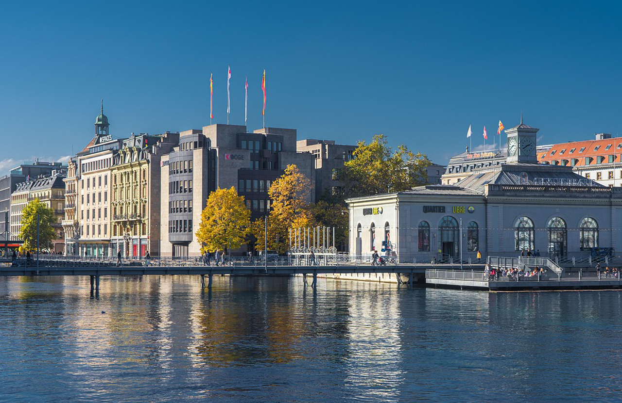 Stadtansicht von Genf mit Gebäuden am Wasser und bunten Bäumen im Vordergrund.