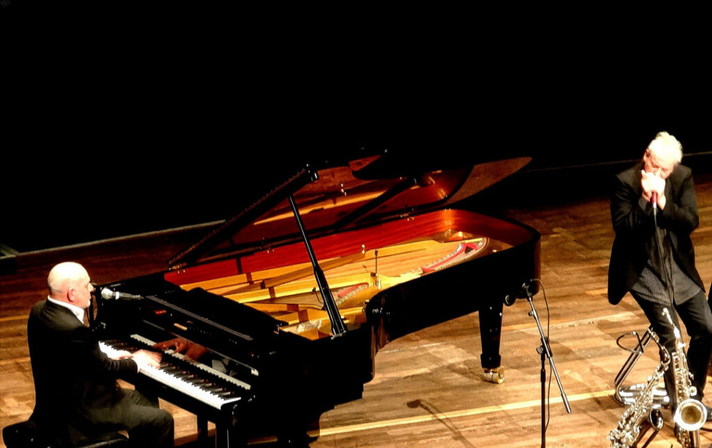 Zwei Musiker auf der Bühne beim Jubiläumskonzert, einer am Klavier, der andere mit Mundharmonika.
