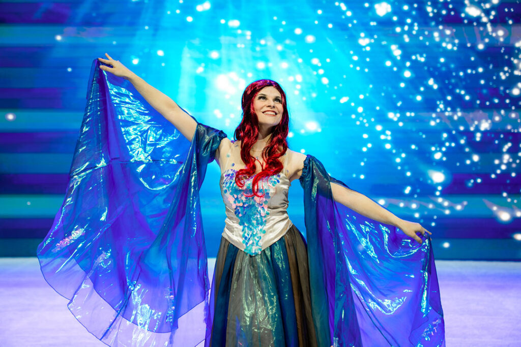Teaserbild zu Musical Dreams mit einer Darstellerin in einem glitzernden Kostüm und blauen Umhängen.