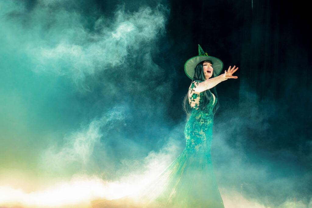 Teaserbild zu Musical Dreams mit einer Sängerin in einem grünen Kleid und Hexenhut auf einer Bühne.
